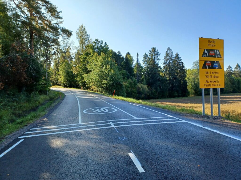 Kuvituskuva, kylätien alku sekä liikennemerkki, joka kuvaa kylätietä. 