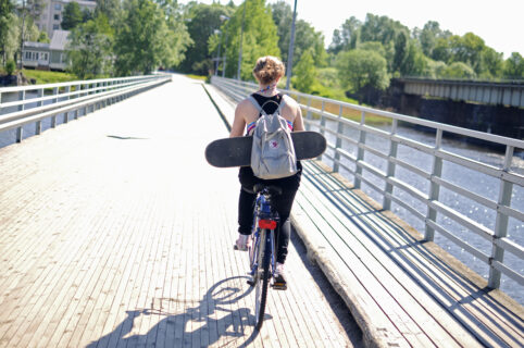 Ung kvinna cyklar över en bro med en skateboard i famnen.