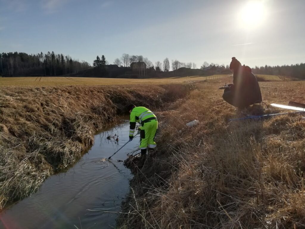 Provtagare samlar ett vattenprov i en vattenfåra i åkerlandskap.